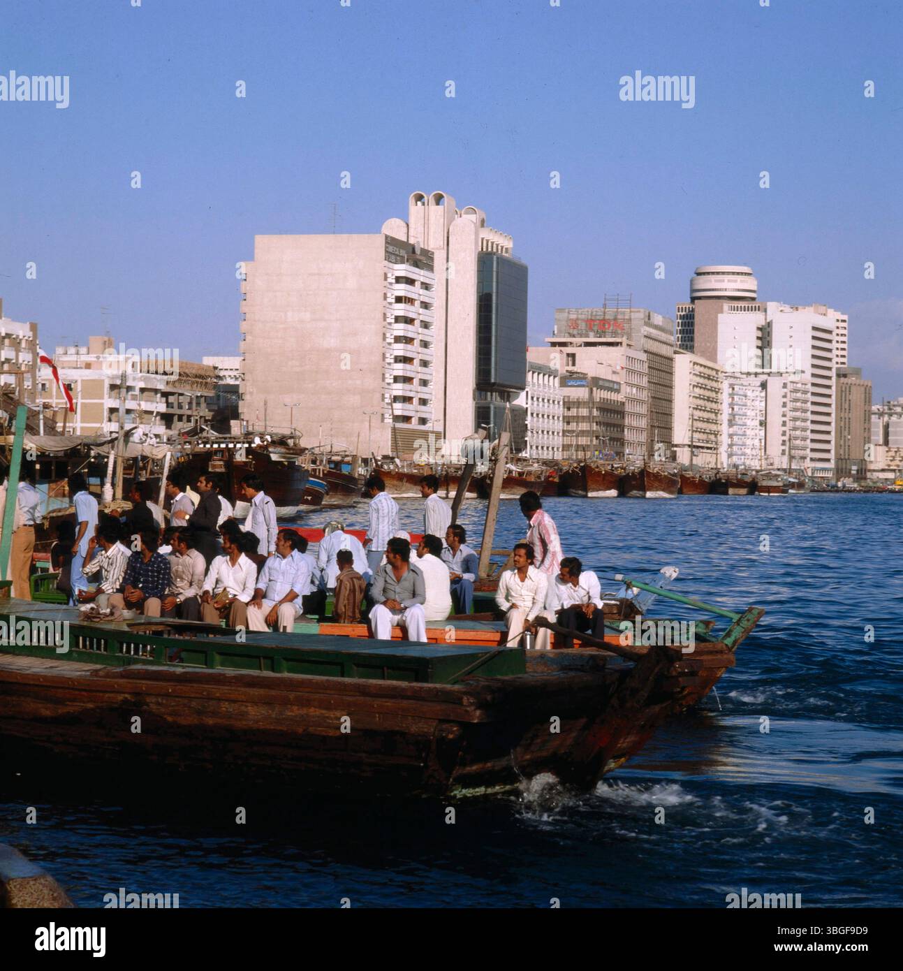 Personengruppe auf einem motorisierten Holzboot auf dem Dubai Creek, mit Blick auf die Uferbebauung des Stadtteils Deira. Im Hintergrund liegen weitere Dhaus entlang des Kais vor einer Reihe von Hochhäusern. Foto Stock