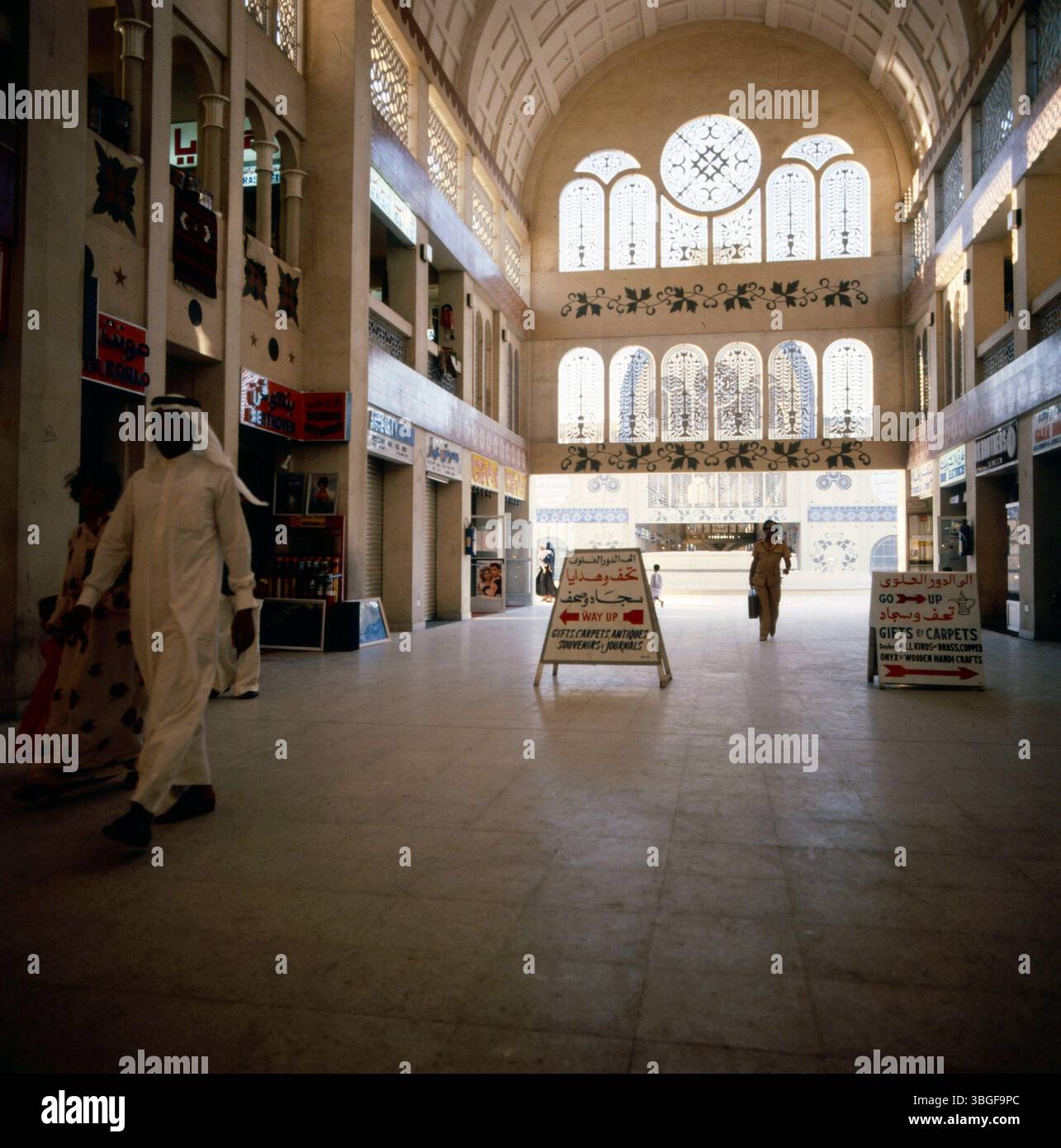 Innenbereich des Sharjah Central Souq mit zweigeschossigen Ladenzeilen, ornamentaler Fensterfassade und Hinweisschildern auf Teppich- und Geschenkartikelgeschäfte. Personen bewegen sich durch die zentrale Halle bei Tageslicht. Foto Stock