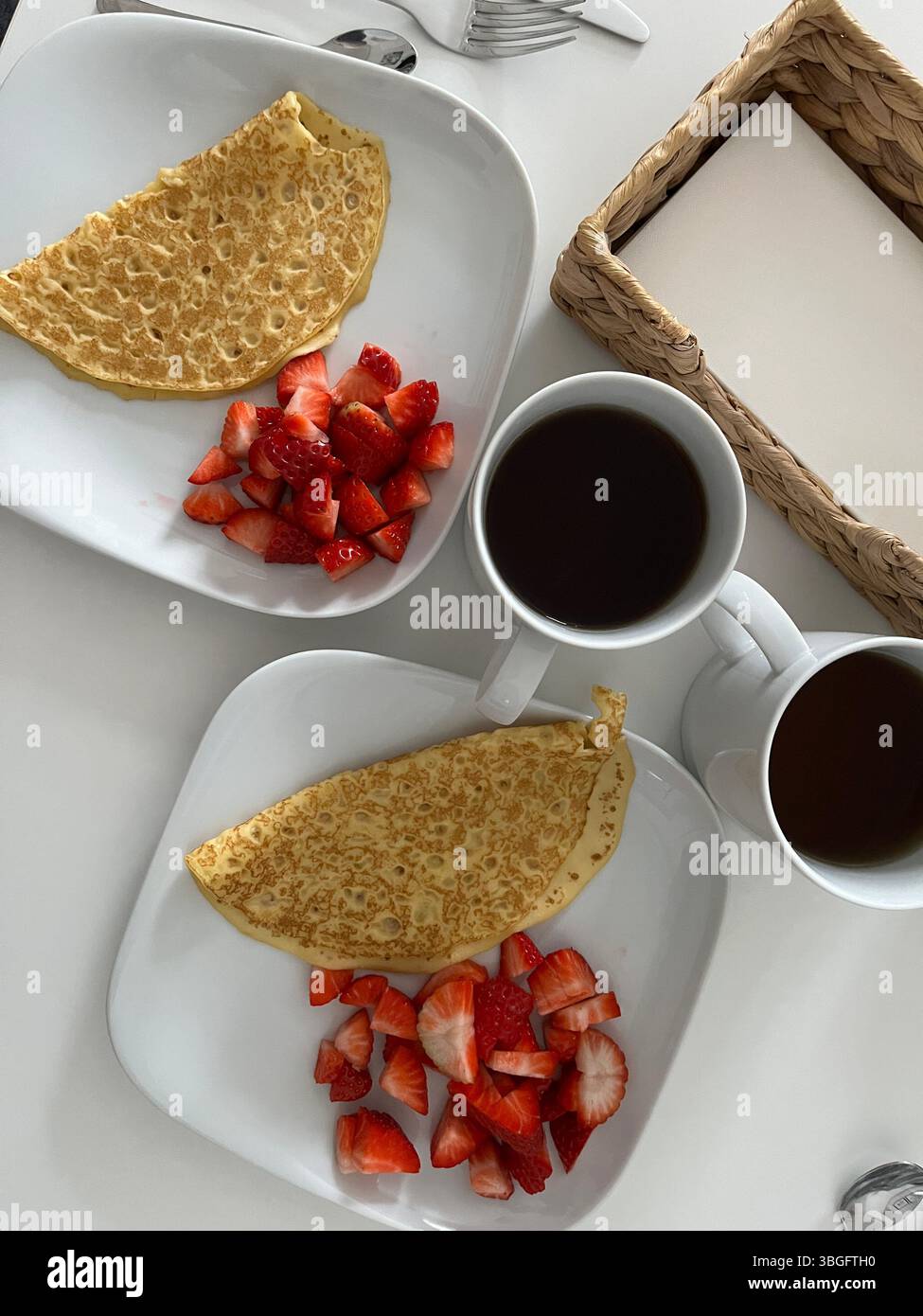 Colazione minimalista con due piatti bianchi di crepes e fragole fresche, serviti insieme a caffè nero in tazze bianche su un tavolo bianco. Foto Stock