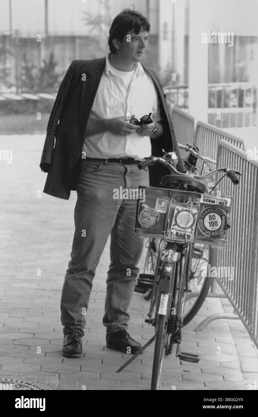 Joschka Fischer, damali hessischer Minister für Umwelt und energie, lehnt am 27. Settembre 1986 un seinem mit Aufklebern beklebten Fahrrad vor einem modernen Regierungsgebäude. Er trägt einen Weißen Hemd, Jeans und ein Jackett Locker über die Schulter geworfen. Die Front Seines Fahrrads ist mit politischen Botschaften versehen – darunter ein Aufkleber mit der Forderung '80 statt 100 – Kampf jetzt gegen das Waldsterben', ein Symbol der Umweltschutzbewegung der 1980er Jahre. Die Aufnahme zeigt Fischer in einem Moment zwischen politischem Proteststil und Ministerrolle. Foto Stock
