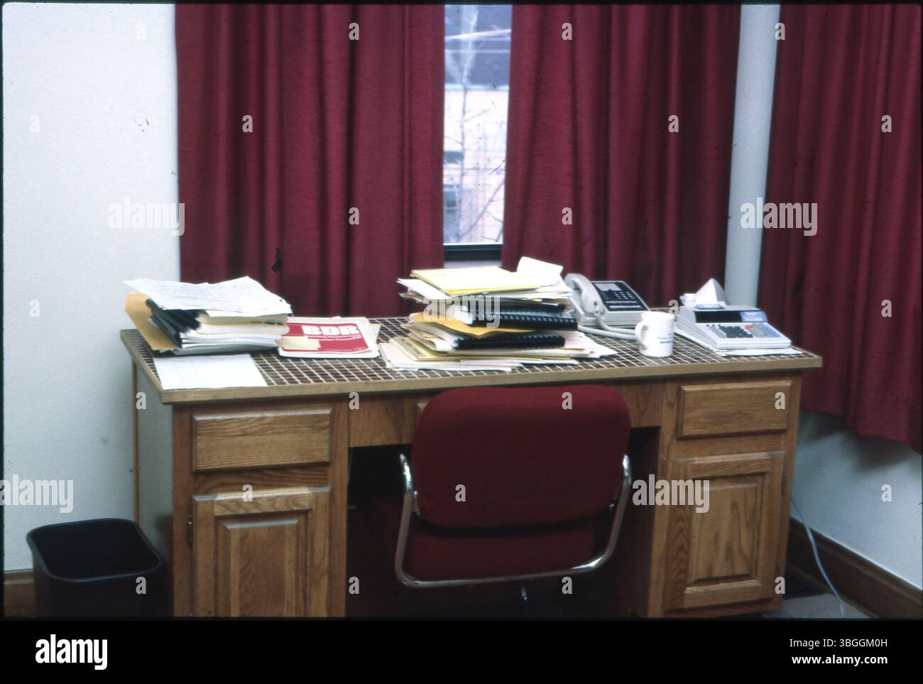 Vista dal 1985 circa di una scrivania da ufficio in legno con pile di carte, una tazza da caffè, un telefono e una macchina aggiuntiva. Le tende rosse sono visibili sullo sfondo. Foto Stock Vista dal 1985 circa di una scrivania da ufficio in legno con pile di carte, una tazza da caffè, un telefono e una macchina aggiuntiva. Le tende rosse sono visibili sullo sfondo. Foto Stock