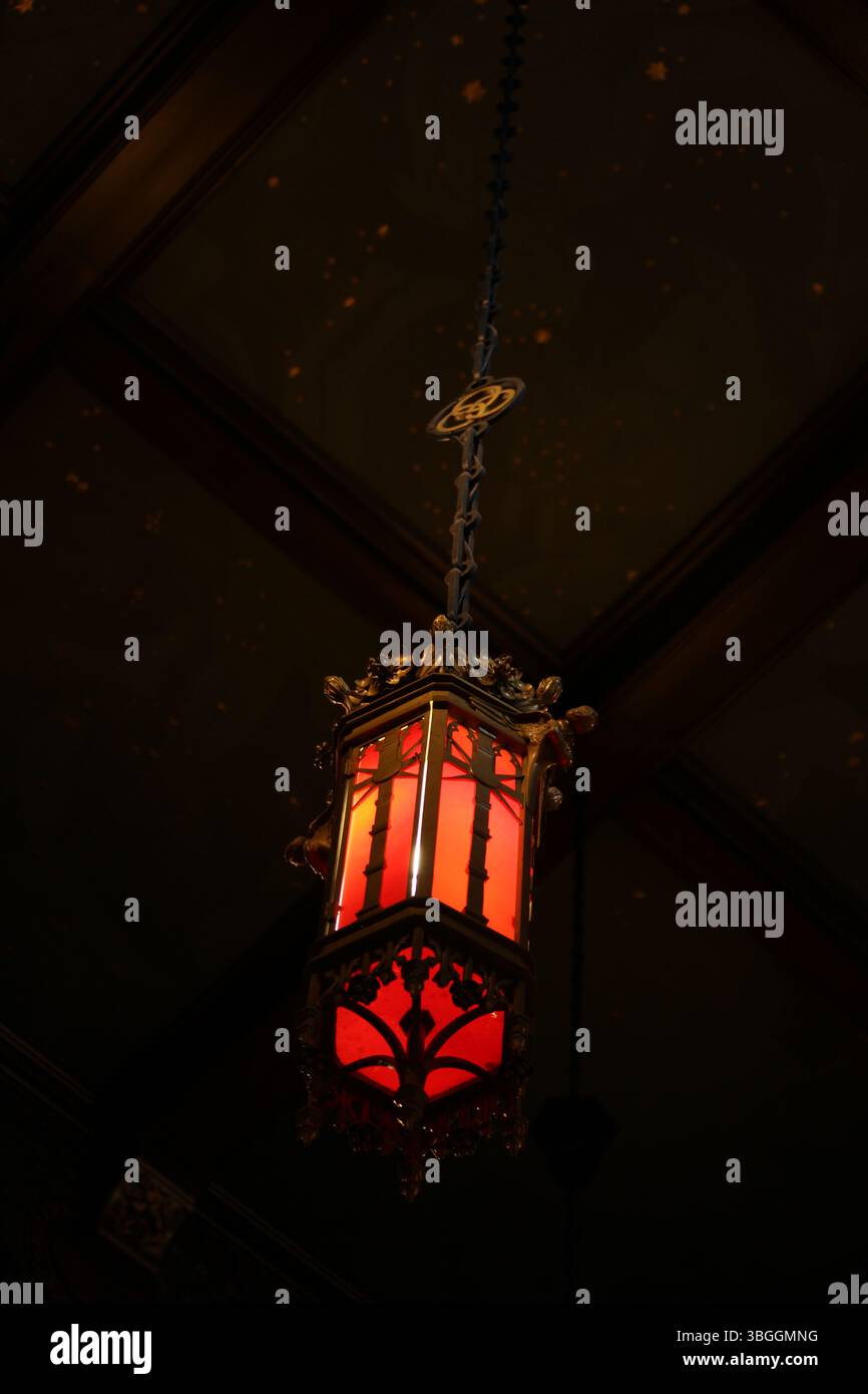 Una lanterna rossa vintage in stile gotico è appesa a una catena su un soffitto scuro e decorato. La lanterna emette un caldo bagliore rosso, creando un misterioso bancomat Foto Stock