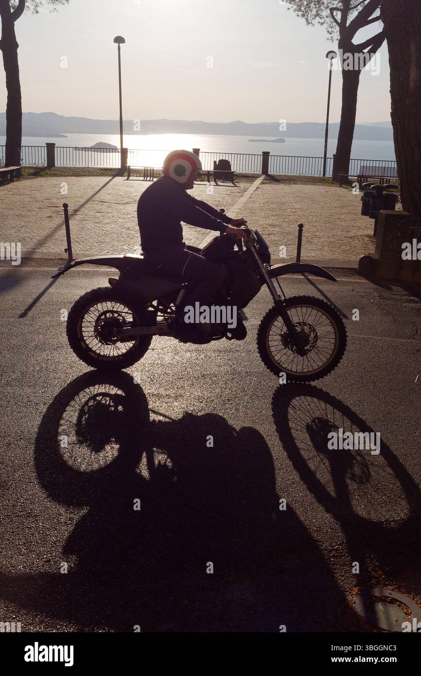 Motociclista con casco casta forte ombra con il lago di Bolsena alle spalle. Montefiascone, regione Lazio, Italia. 3 giugno 2025 Foto Stock