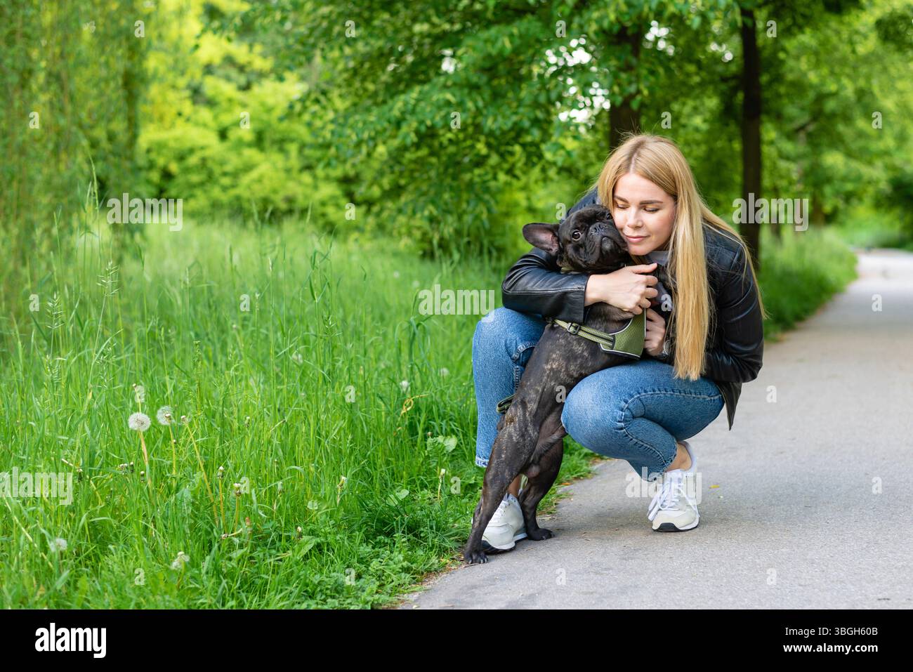 Una giovane donna bionda che abbraccia dolcemente un bulldog francese nero in un parco verde. Il cane guarda direttamente la fotocamera con un'espressione seria mentre è in vita Foto Stock Una giovane donna bionda che abbraccia dolcemente un bulldog francese nero in un parco verde. Il cane guarda direttamente la fotocamera con un'espressione seria mentre è in vita Foto Stock