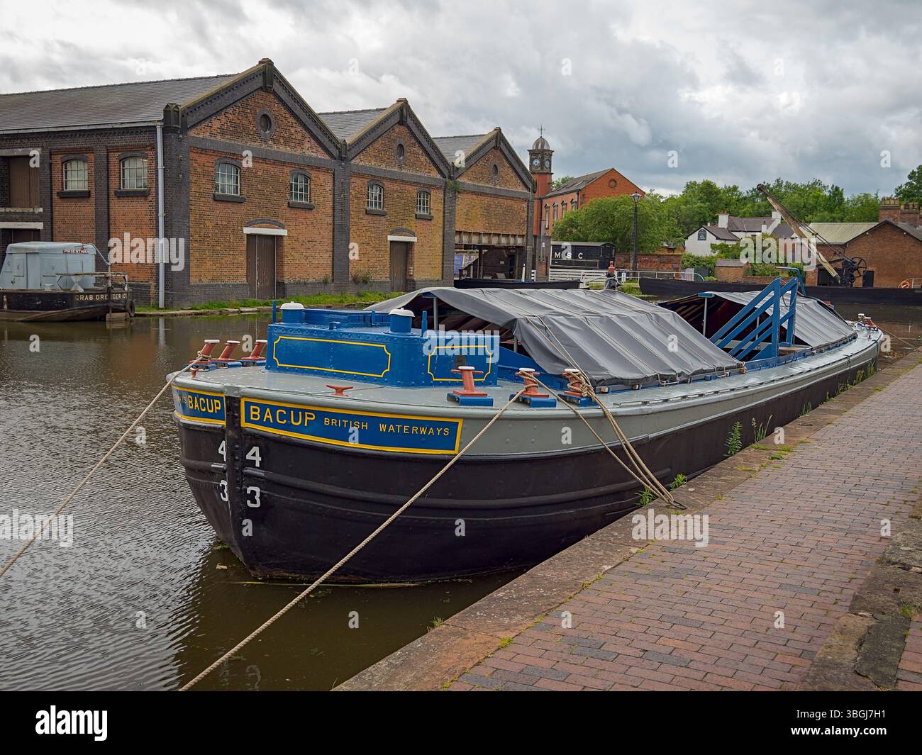 Ellesmere Port, Cheshire, Regno Unito, 05-31-2025: Museo nazionale dei corsi d'acqua. Storico battello sul canale attraccato da magazzini in mattoni d'epoca sotto un cielo nuvoloso. Foto Stock