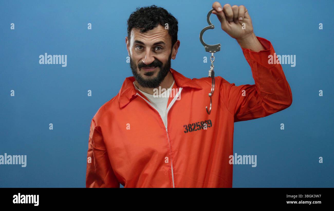 Uomo ispanico con barba in tuta da prigione arancione che tiene le manette su sfondo blu, raffigurante il tema dell'incarcerazione. Foto Stock