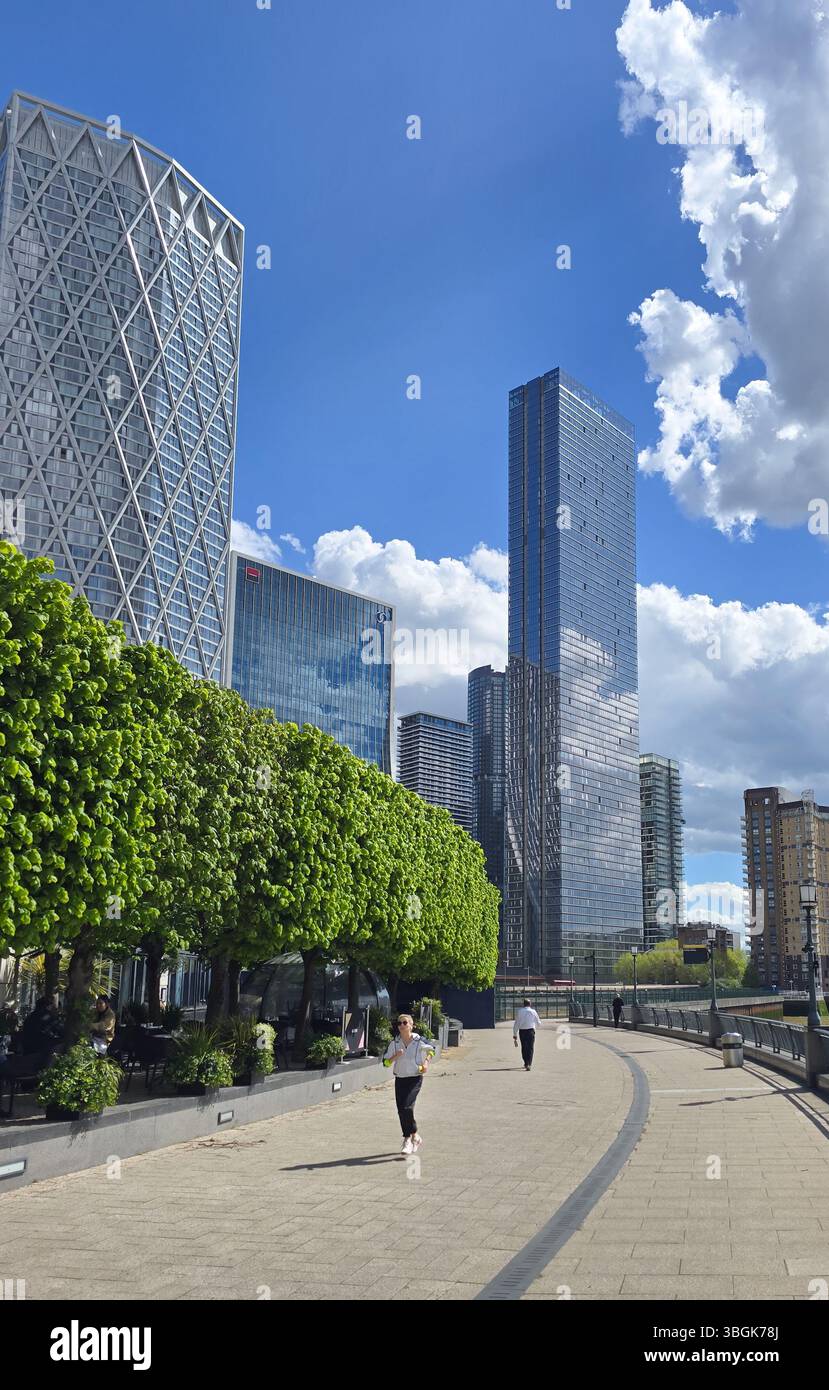 Londra, Regno Unito. 17 aprile 2025, persone che camminano e fanno jogging a Canary Wharf, Londra, con moderni grattacieli e alberi verdi sotto un cielo blu con nuvole Foto Stock