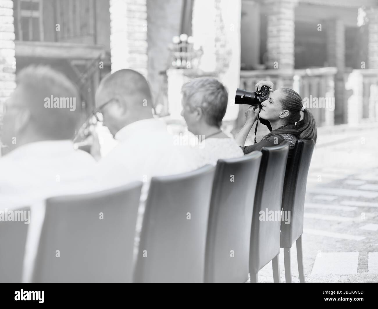 Fotografia in bianco e nero di una donna con una fotocamera all'aperto accanto a un pubblico seduto durante un evento. Foto Stock