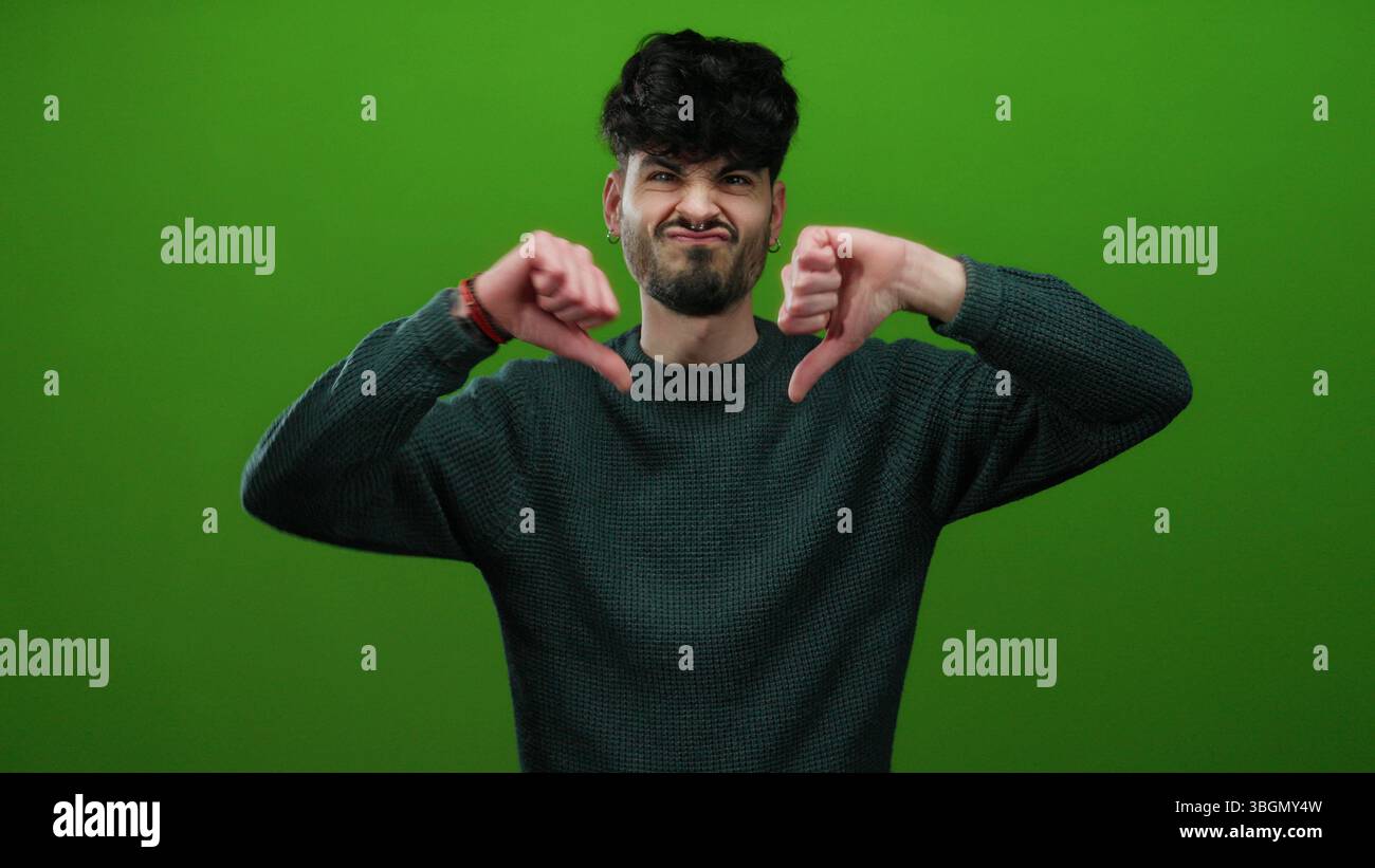 Uomo che esprime disprezzo con un gesto verso il basso contro il muro verde in un ambiente isolato, che ritrae vividamente disapprovazione. Foto Stock