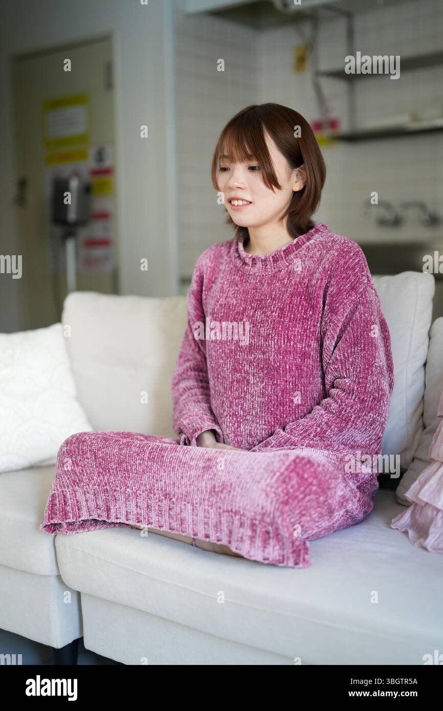 In una stanza tranquilla e ben illuminata, una donna giapponese incinta di vent'anni con i capelli marroni corti, che indossa un abito a maglia rosa, siede su un divano per esercitarsi Foto Stock