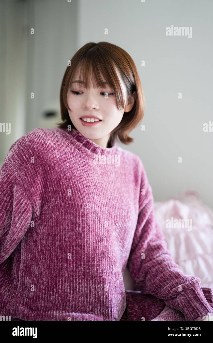 In una stanza tranquilla e ben illuminata, una donna giapponese incinta di vent'anni con i capelli marroni corti, che indossa un abito a maglia rosa, siede su un divano per esercitarsi Foto Stock