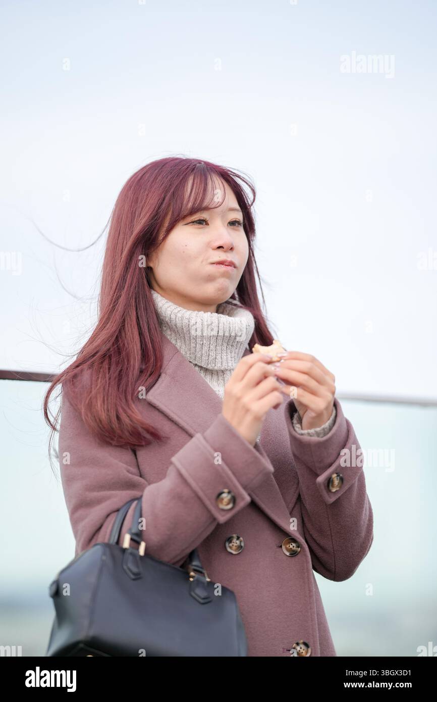 In una fredda mattina di dicembre, una donna giapponese di vent'anni con lunghi capelli rosa e un trench rosa si erge su un tetto sopra la città. Sorridere, Foto Stock
