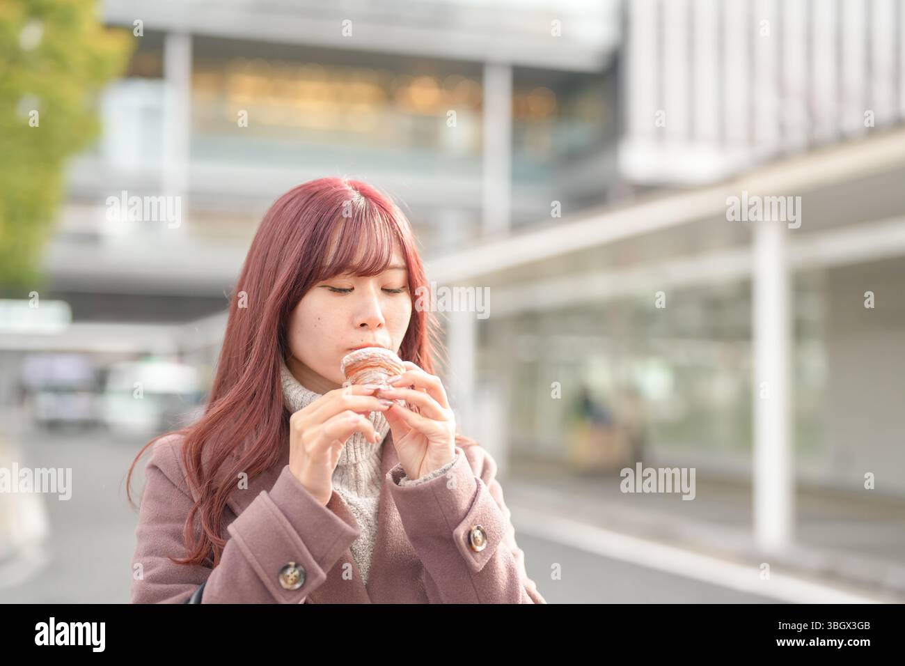 In una fredda mattina di dicembre, una donna giapponese ventenne con lunghi capelli rosa, con indosso un trench rosa, mangia felicemente un croissant davanti a H. Foto Stock