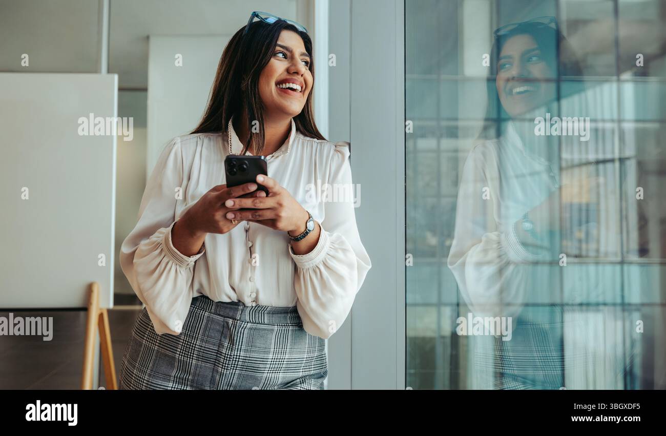 Una giovane donna professionale si trova accanto a una luminosa finestra di ufficio, tenendo in mano uno smartphone e sorridendo. La sua espressione riflette fiducia e successo, Highlig Foto Stock