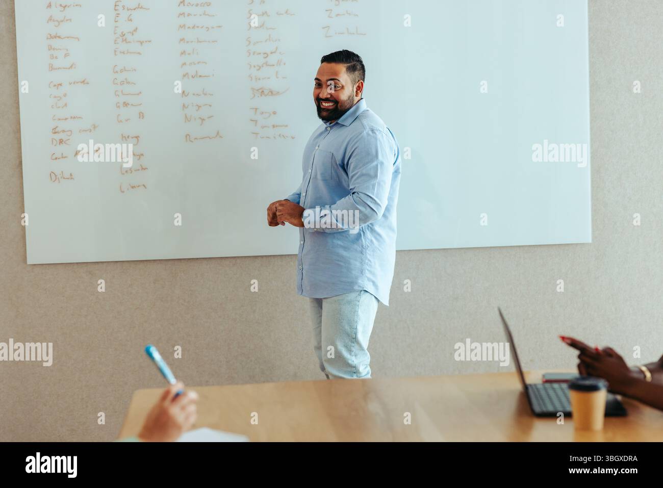 Un allegro uomo d'affari sta presentando idee su una lavagna durante una riunione in un ufficio moderno. I colleghi ascoltano, con uno che prende appunti Foto Stock