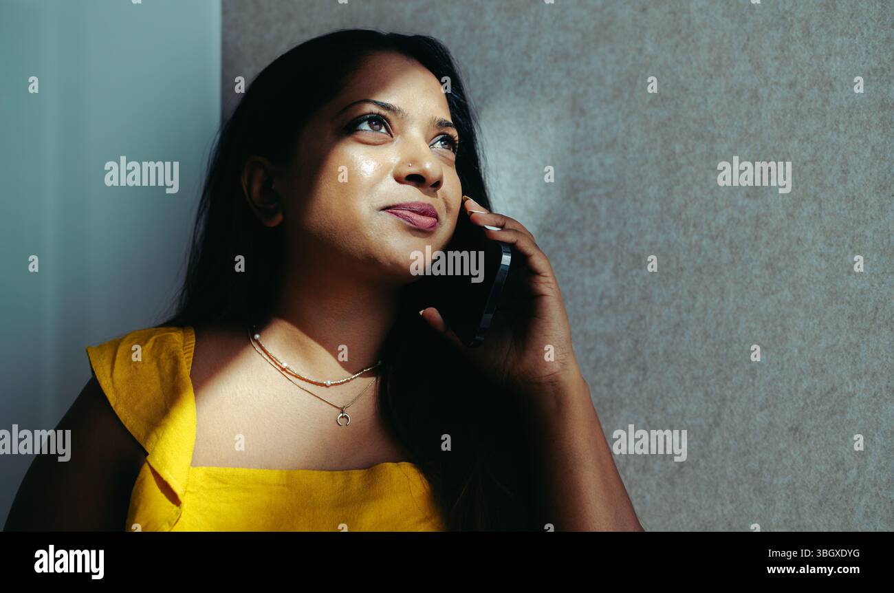 Donna d'affari indiana che parla sul suo telefono cellulare, accanto a una parete illuminata. La sua espressione trasmette concentrazione e determinazione mentre ingerisce Foto Stock