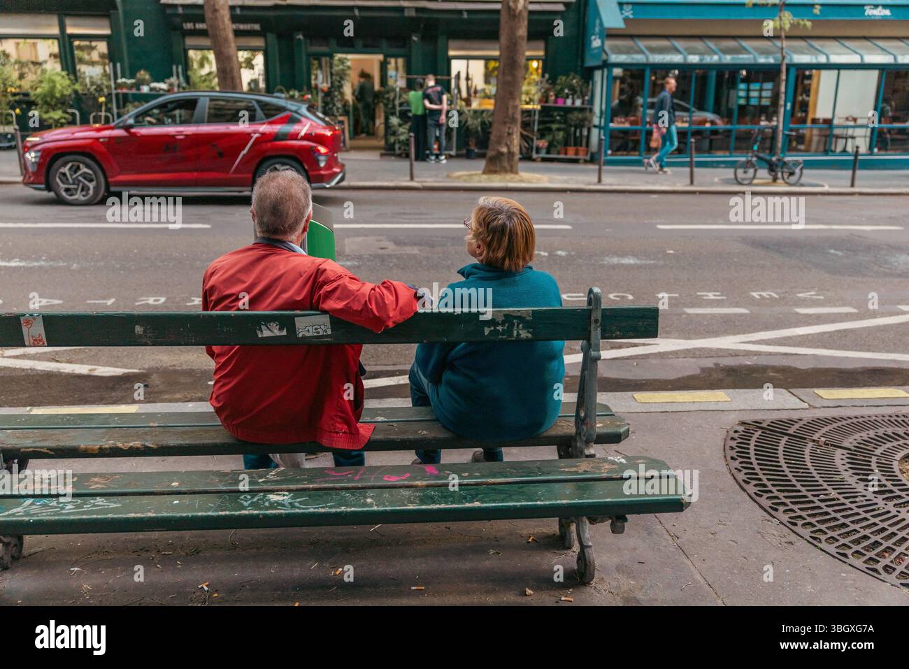 Una coppia di anziani, vista da dietro, siede da vicino su una panchina verde, condividendo un momento tranquillo mentre si affacciano su una vivace strada della città. Il loro postu Foto Stock