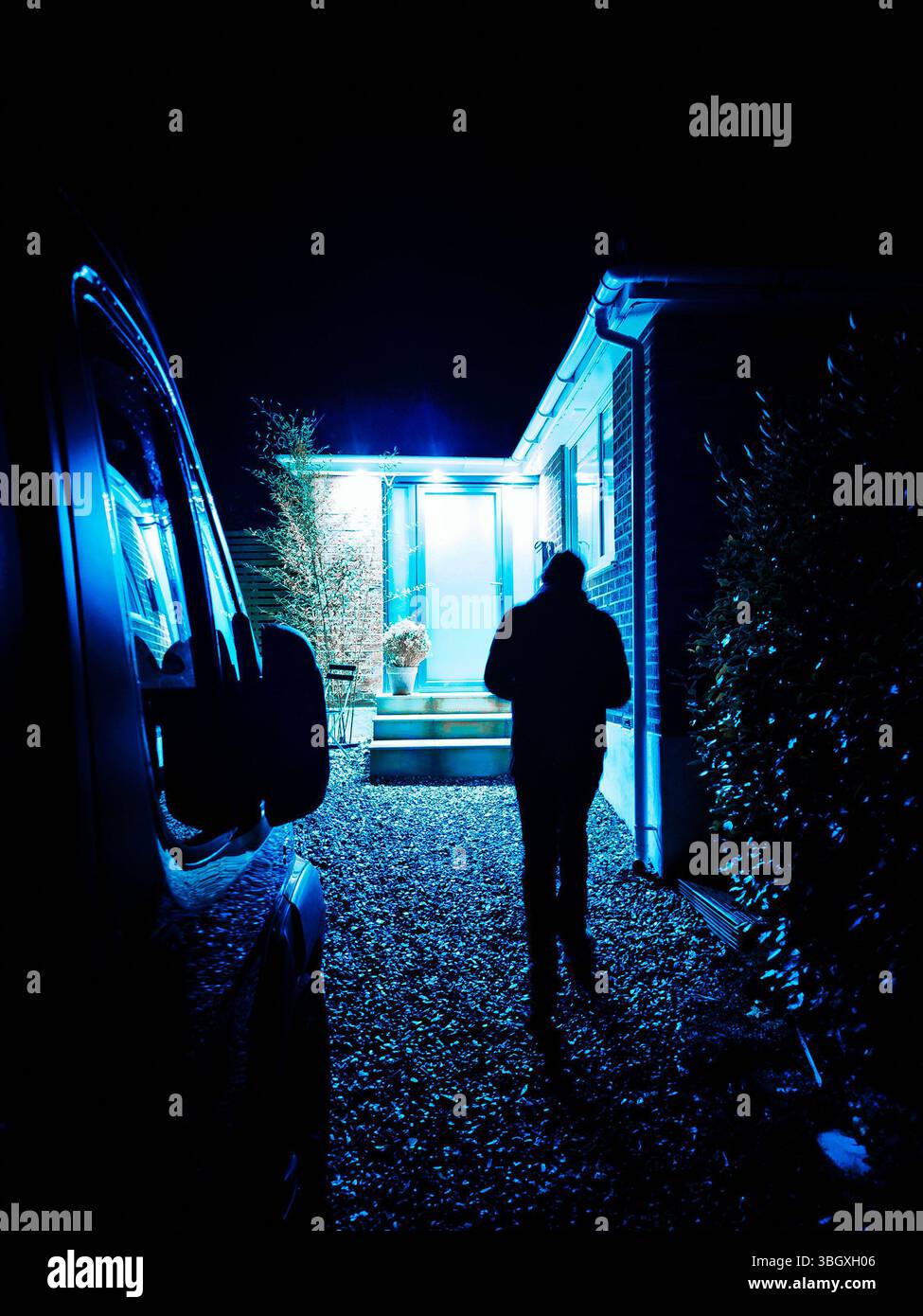 Sagoma di una persona che cammina verso un ingresso luminoso di notte in una casa moderna, con illuminazione blu, copertina del libro thriller con furto Foto Stock