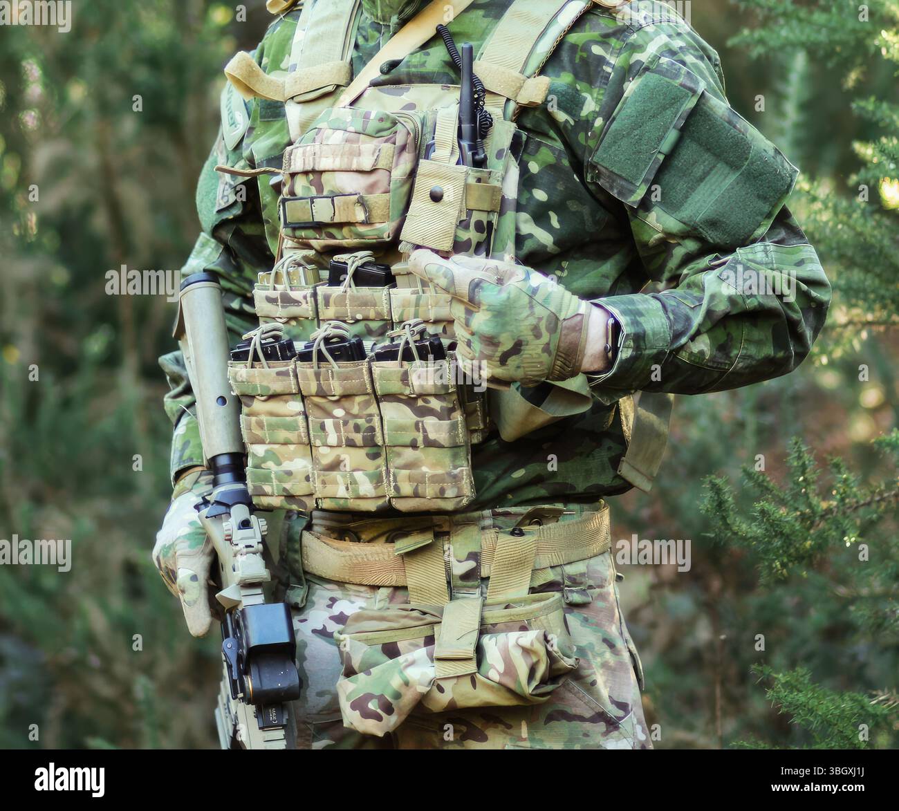 Soldato in uniforme mimetica e giubbotto tattico che punta su riviste di munizioni durante l'esercitazione di combattimento in un'area boscosa, dimostrando prontezza e preparazione al combattimento Foto Stock