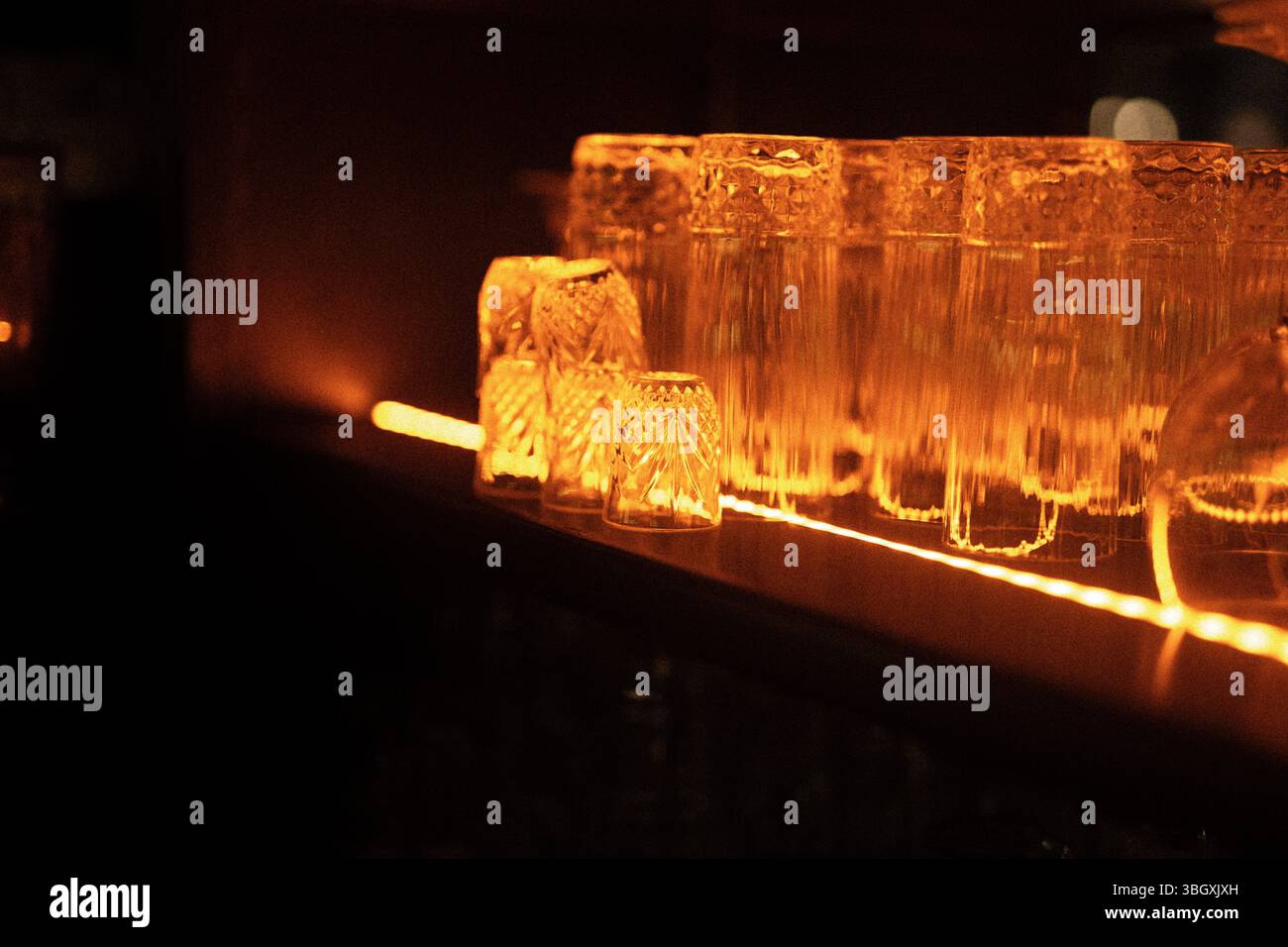 In un bar buio e dall'atmosfera suggestiva, una fila di bicchieri puliti si trova su uno scaffale, che si illumina dalla calda luce arancione di una striscia a LED sottostante. Foto Stock
