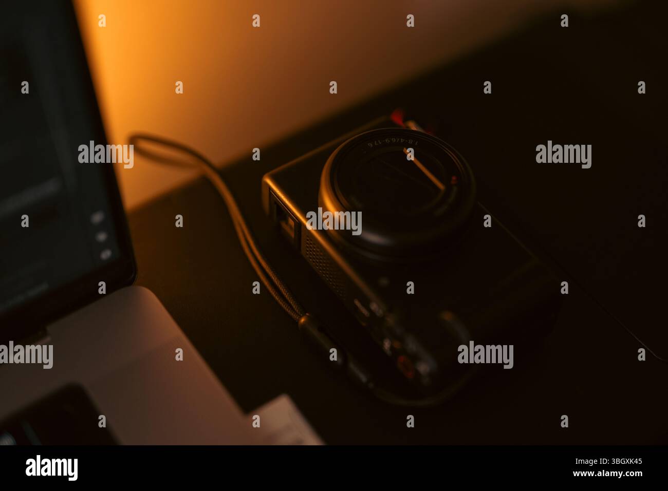 Una fotocamera digitale compatta, elegante e nera, si appoggia su una superficie scura, illuminata da un'illuminazione calda e suggestiva. Un notebook è parzialmente visibile nel backgro Foto Stock