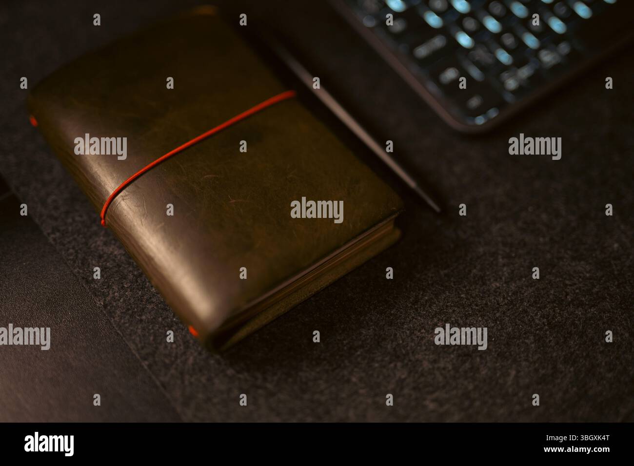 Un notebook marrone rilegato in pelle con fascia elastica arancione e penna si trova su una superficie scura e ruvida, sotto una luce soffusa e calda, che evoca un senso di quie Foto Stock