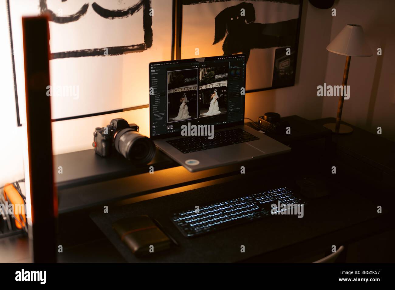 La scrivania di un fotografo è poco illuminata e include un laptop che visualizza le foto di un matrimonio in fase di editing, una fotocamera digitale con obiettivo, una tastiera esterna, una Foto Stock