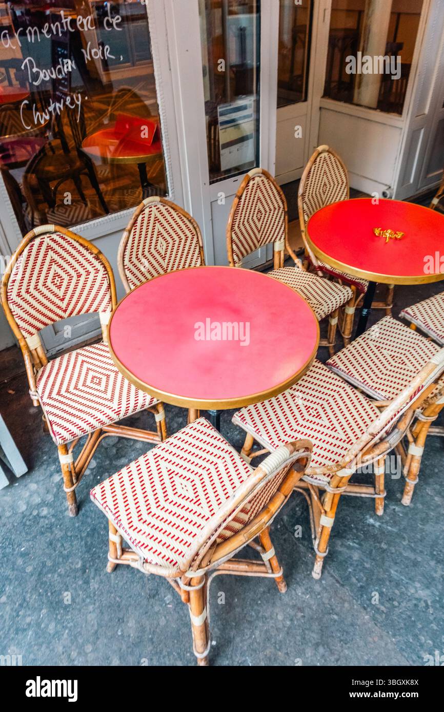 Sedie vuote e vivaci in tessuto rosso e bianco sono disposte intorno a tavoli rotondi di rosso luminoso sulla terrazza di un caffè parigino. La scena evoca questo Foto Stock