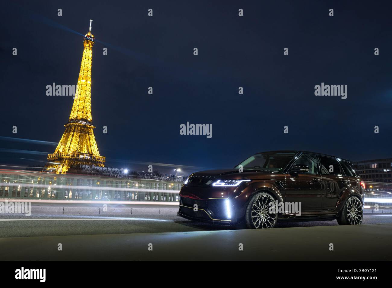 Un SUV di lusso dai colori scuri è parcheggiato in una strada di notte, con sullo sfondo l'iconica torre di Parigi illuminata brillantemente. Sentieri leggeri da Foto Stock