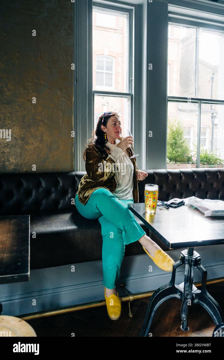 Donna che sorseggia del vino in un'accogliente sala da pub vicino a una finestra soleggiata Foto Stock
