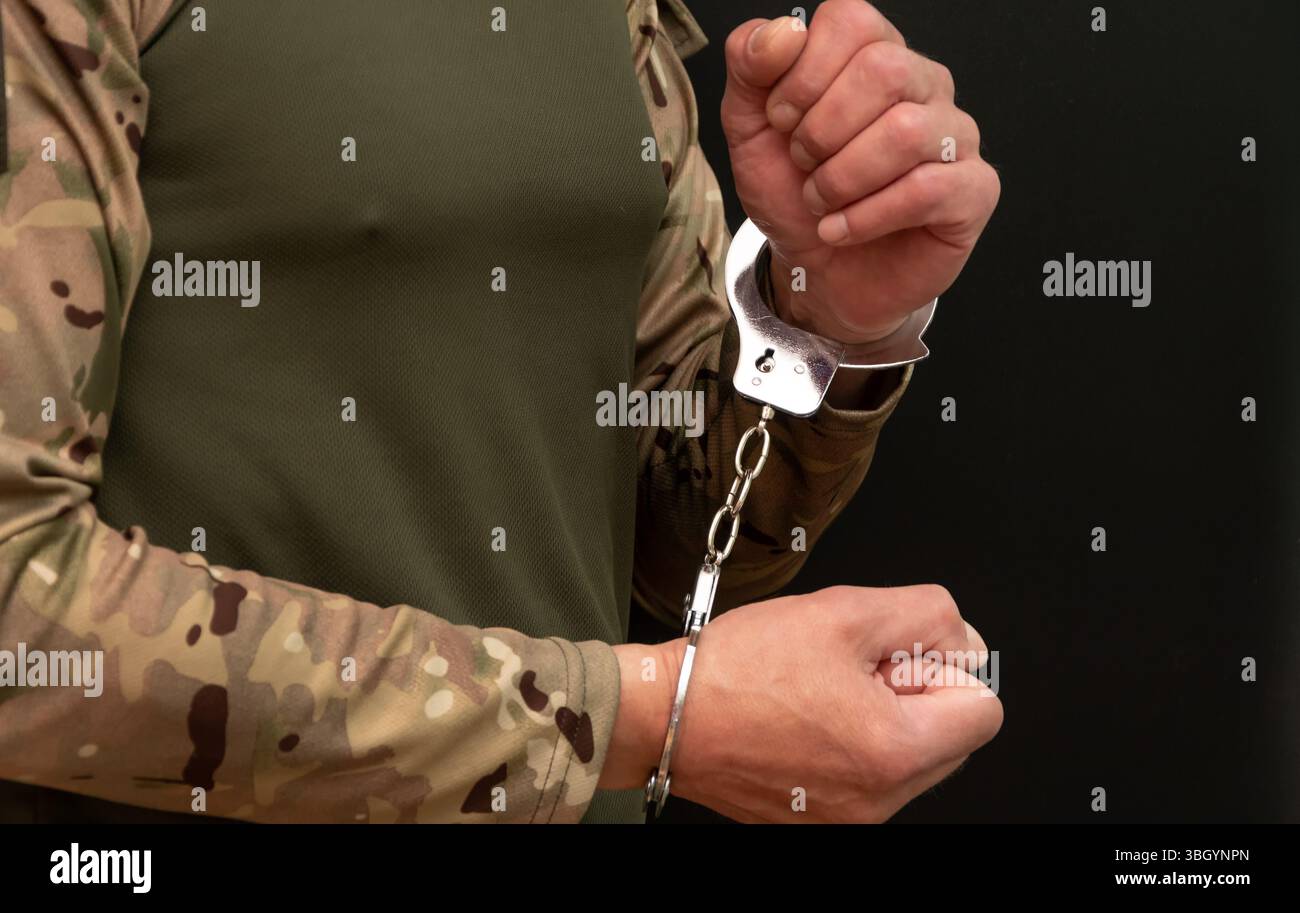 Primo piano di mani ammanettate di un uomo in abiti militari su uno sfondo scuro Foto Stock