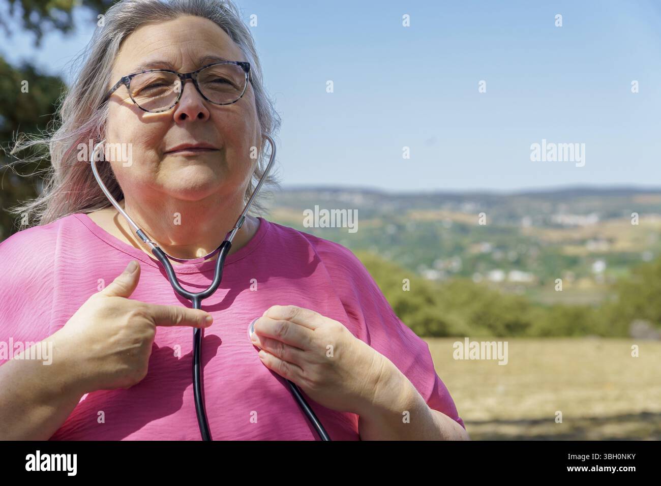 Donna matura che usa uno stetoscopio, esegue un autocontrollo sulla sua salute del cuore, promuovendo un invecchiamento sano e l'autoassistenza Foto Stock