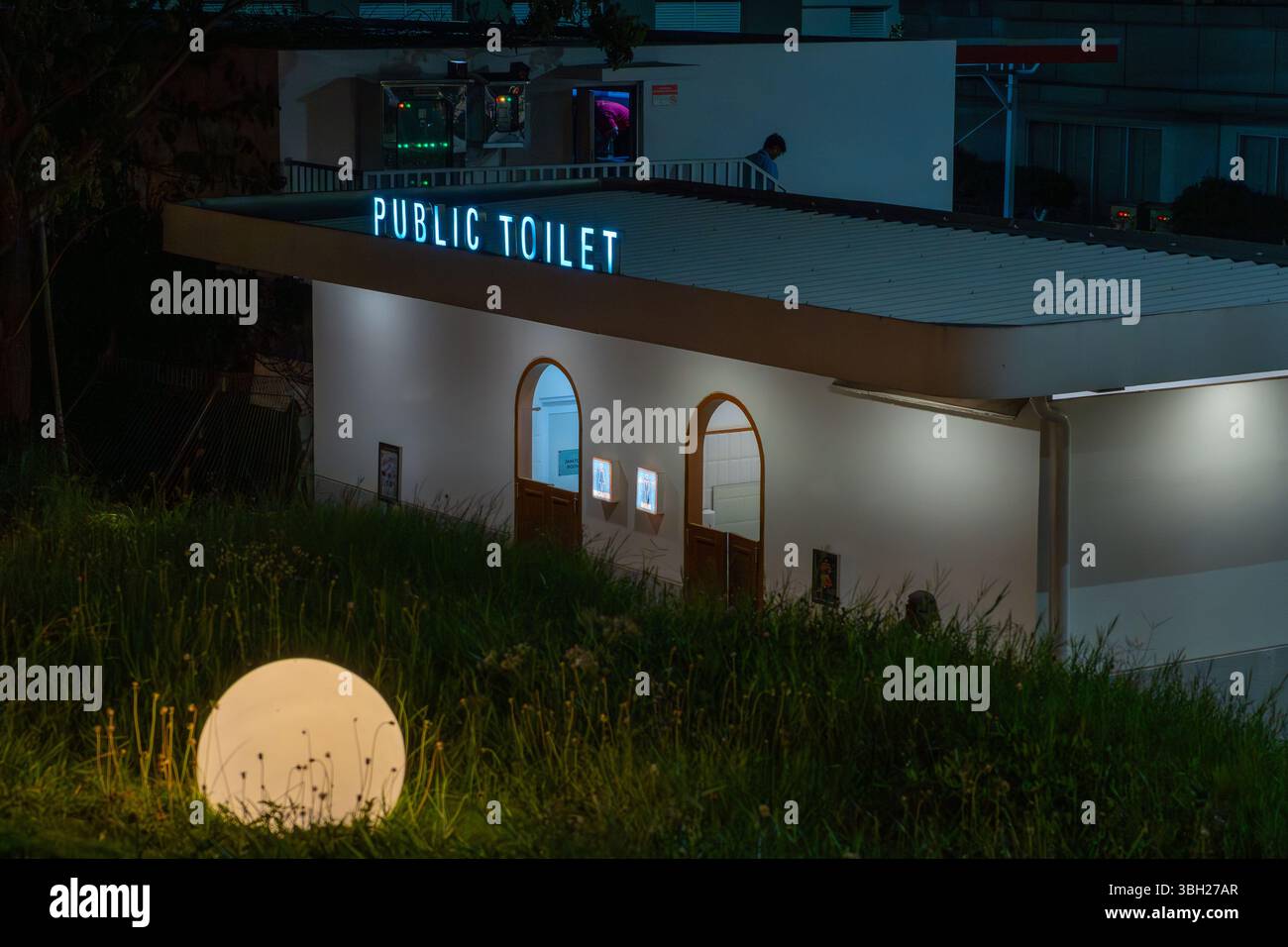 Edificio del bagno pubblico con un tetto bianco e un cartello blu che dice "servizi igienici pubblici". L'edificio è illuminato di notte Foto Stock