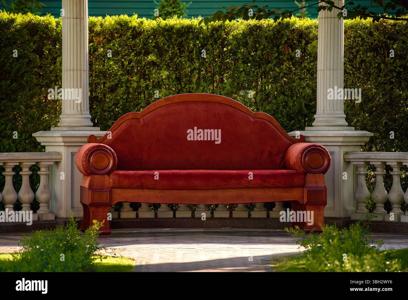 Lussuoso divano rosso in stile vintage con dettagli in legno collocato in un classico padiglione giardino. Circondato da colonne bianche, balaustre e vegetazione. Foto Stock