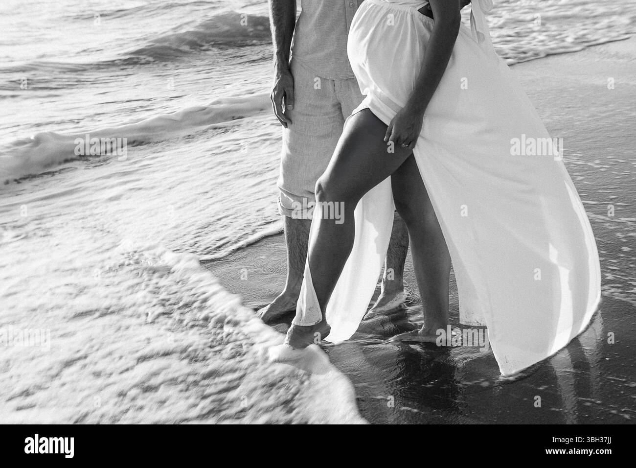 Una coppia gode di una tranquilla passeggiata lungo la spiaggia mentre le onde si infrangono dolcemente ai piedi. L'immagine cattura la tenerezza di una coppia che si aspetta assaporando un p Foto Stock
