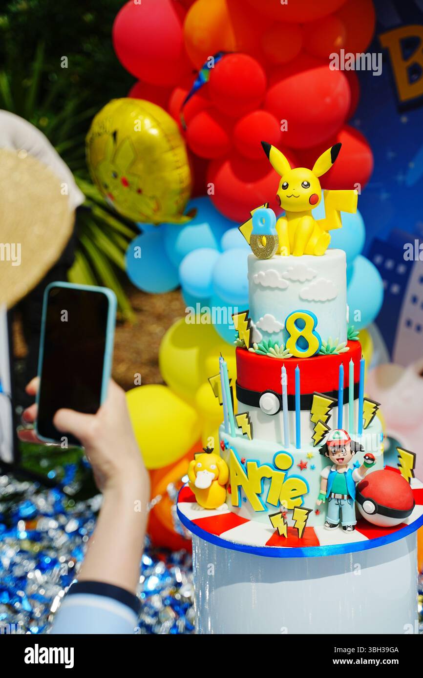 Uno smartphone cattura una vivace torta di compleanno a tema Pokemon adornata con Pikachu e decorazioni colorate. La scena mostra festeggiamenti gioiosi vib Foto Stock