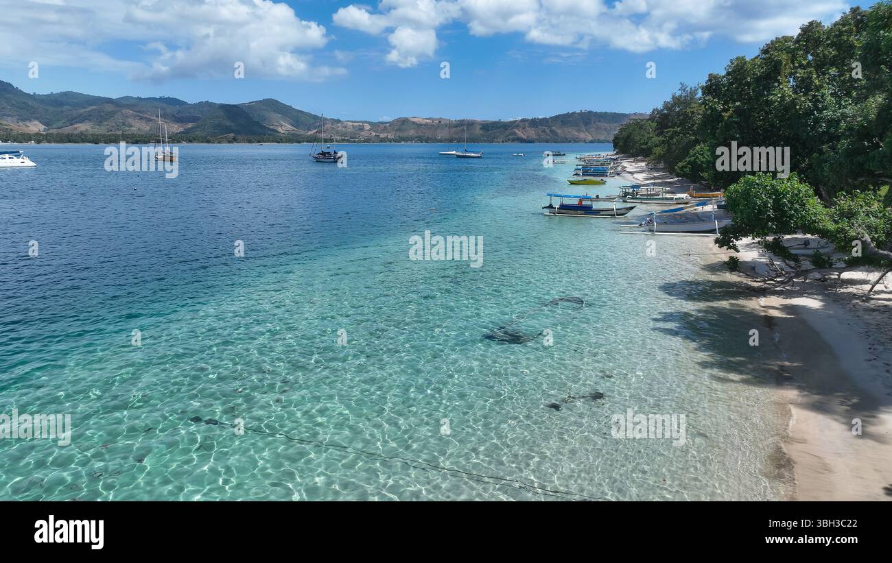 L'acqua limpida e poco profonda rivela il fondale sabbioso lungo una spiaggia tranquilla punteggiata di barche, con montagne panoramiche che si innalzano sullo sfondo sotto il cielo soleggiato Foto Stock