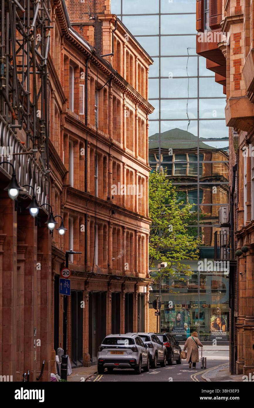 Una strada stretta nel centro di Londra fiancheggiata da edifici in mattoni rossi, auto parcheggiate e una moderna facciata in vetro che riflette l'architettura e il verde Foto Stock