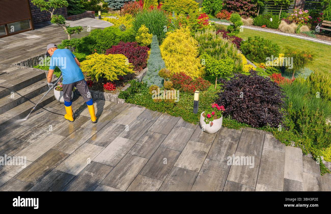 Un giardiniere con stivali gialli innaffia fiori e vegetazione vivaci in uno splendido giardino sul retro, creando un'atmosfera tranquilla e invitante. Foto Stock