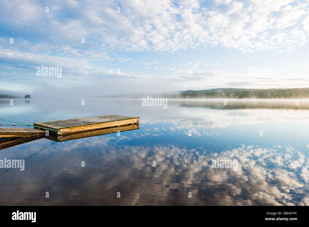 Un molo di legno si estende in un tranquillo lago in Svezia, circondato da nebbie mattutine e morbide nuvole. L'acqua riflette il cielo vibrante, creando un ambiente tranquillo Foto Stock