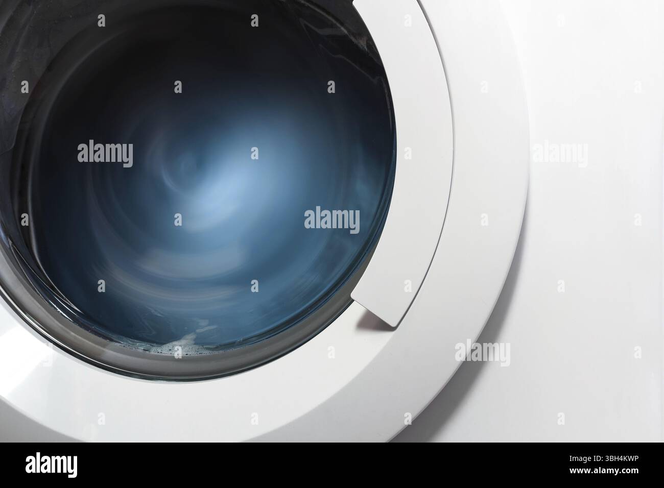 Vista frontale e la porzione dell'oblò della macchina di lavaggio durante il lavaggio e la rotazione del cestello contenente i vestiti blu Foto Stock