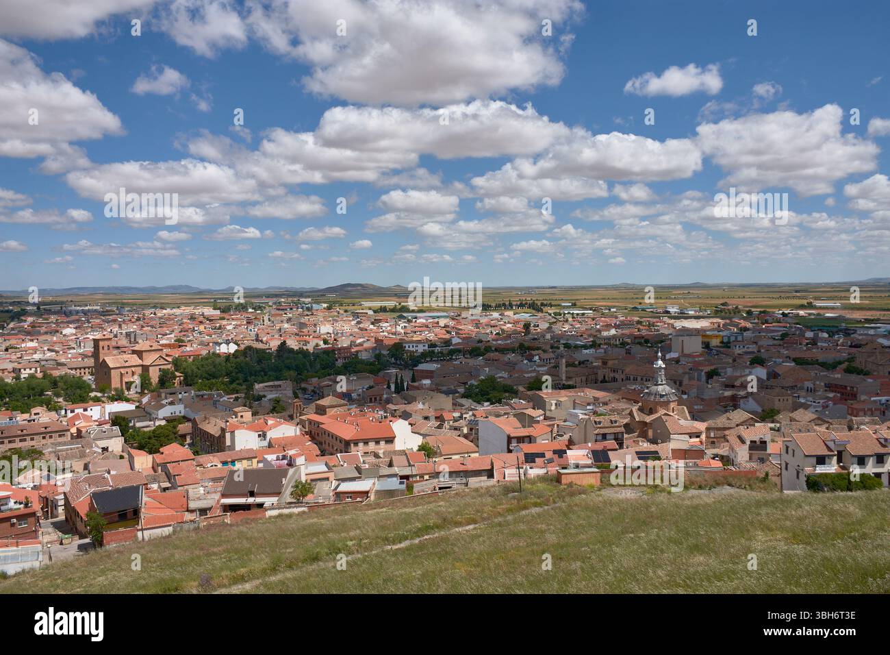 Una vista grandangolare della città di Consuegra, Toledo, mostra tetti di tegole rosse, un'importante chiesa storica e terreni agricoli circostanti sotto un blu brillante Foto Stock