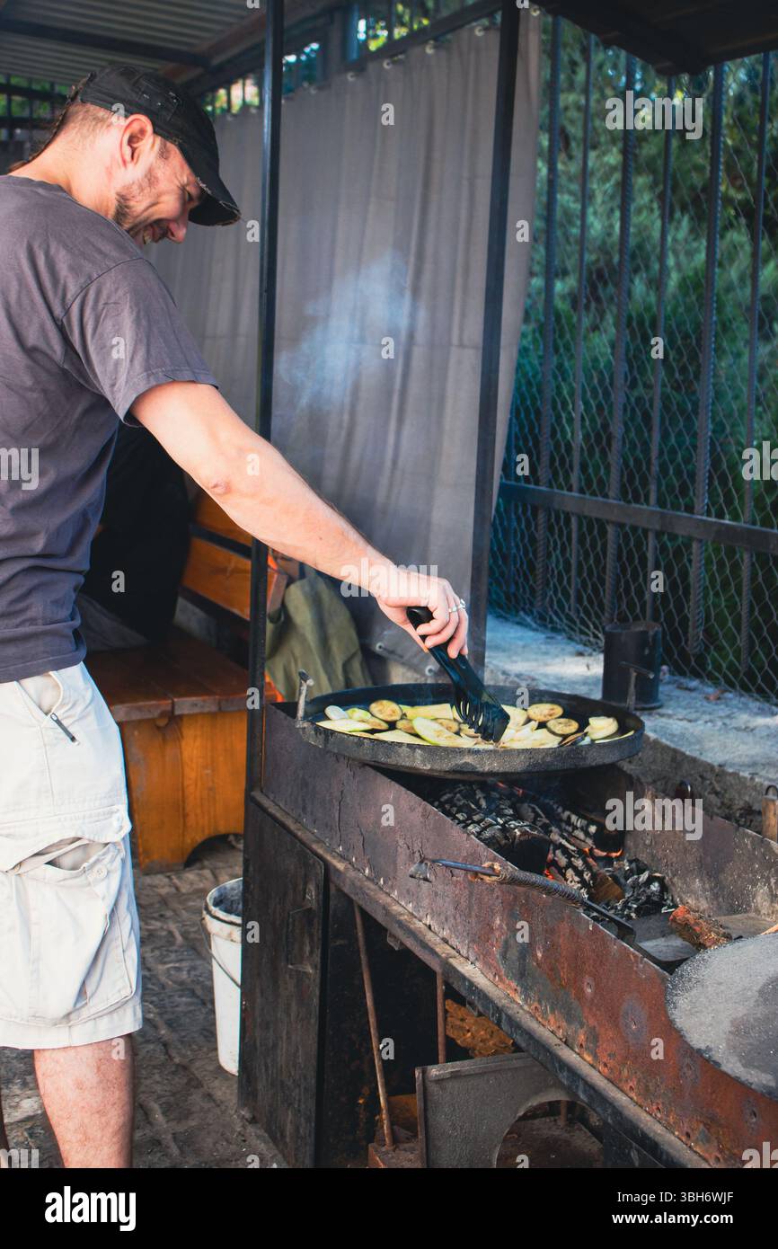 Uomo in poltrona che grigia verdure nel patio. Uomo sorridente al barbecue sul cortile. Attività estive. Carne e verdura nel wok durante la cottura al falò Foto Stock