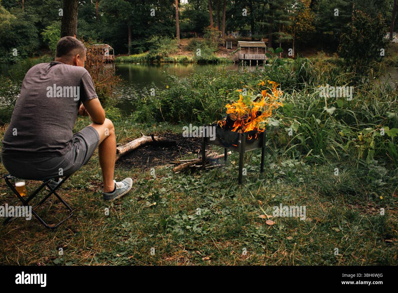 Uomo seduto su una sedia che grigia sulla riva del lago. Uomo al barbecue nella foresta estiva. Attività estive. Festa al barbecue in estate Foto Stock