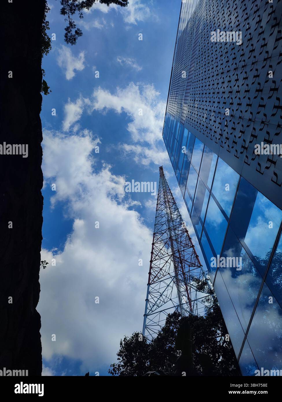 Torre di telecomunicazione in metallo accanto a un edificio riflettente sotto un cielo blu con nuvole: L'industria incontra il design urbano. - Immagine stock catturata con smartphone