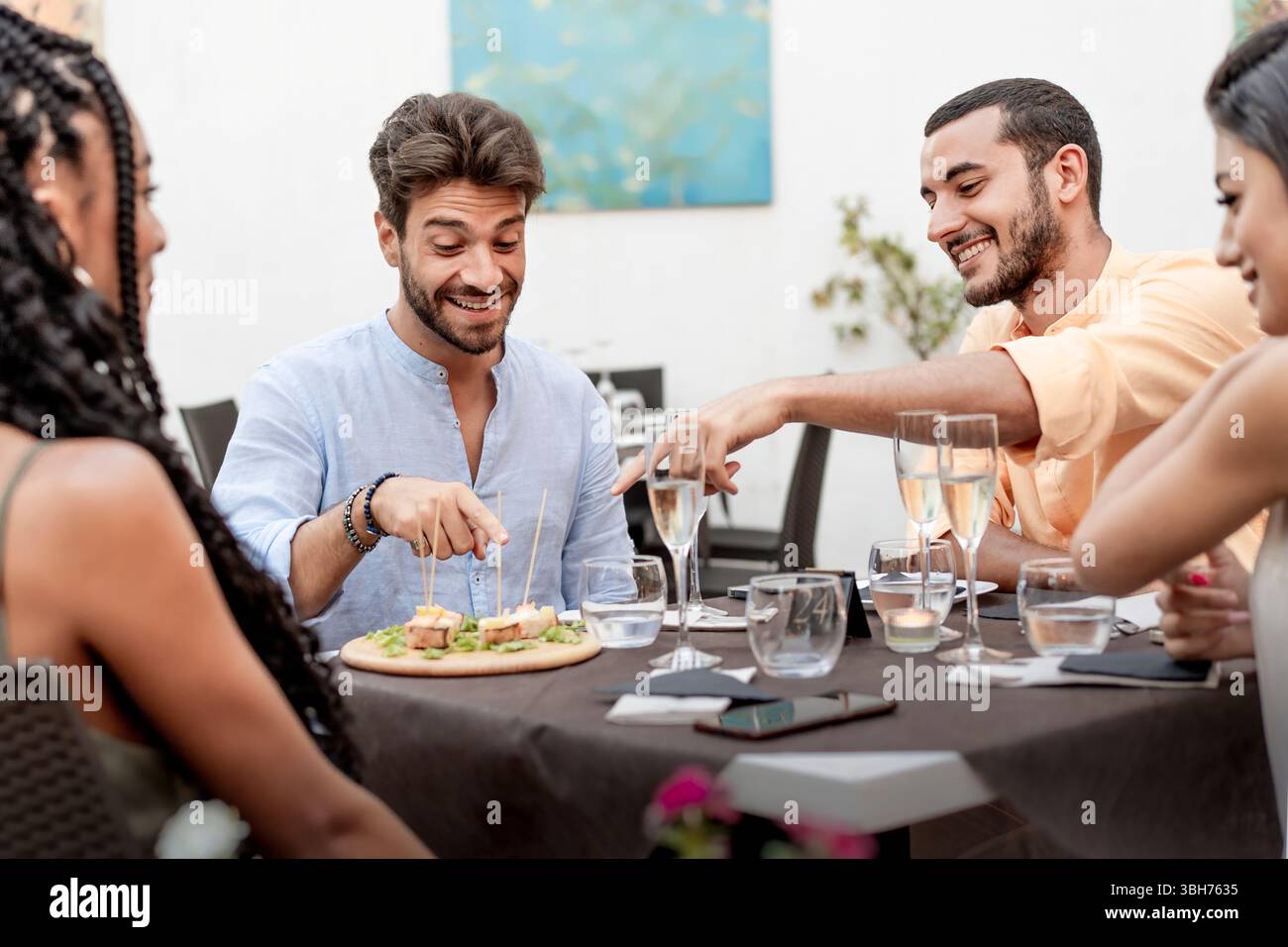 Un gruppo variegato di giovani amici felici condivide antipasti e spumante, socializzando in un elegante ristorante o bar. Una gioiosa scena di amicizia Foto Stock