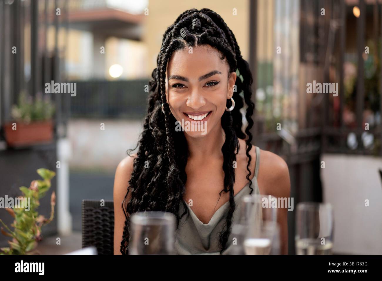 Una giovane donna di colore bella e sicura di sé con un'acconciatura intrecciata sorride calorosamente alla macchina fotografica mentre si siede in un ristorante o in un bar all'aperto. A. Foto Stock