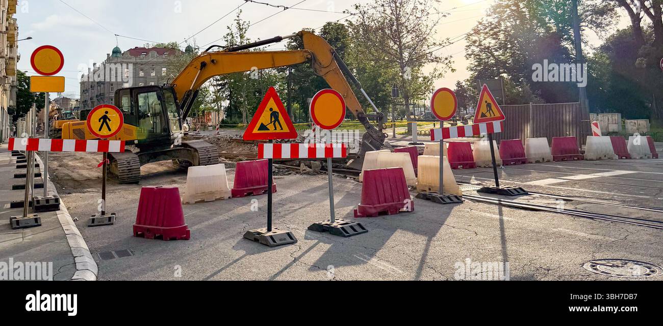 Striscione di cantiere urbano con escavatore e segnali di avvertimento durante le riparazioni stradali. Infrastrutture urbane e lavori pubblici Foto Stock