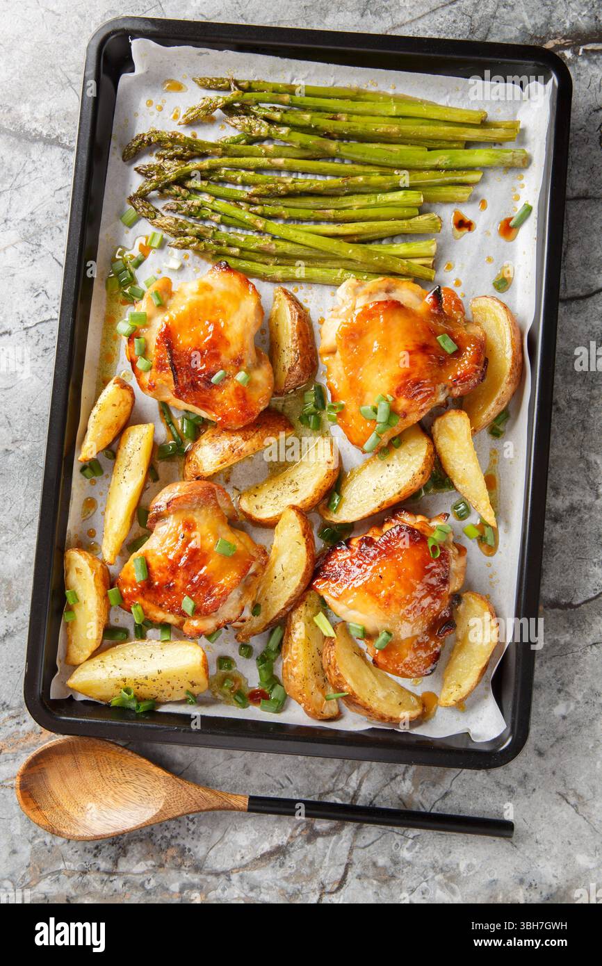Cosce di pollo al forno con spicchi di patate e asparagi in primo piano su una teglia da forno sul tavolo. Vista dall'alto verticale Foto Stock