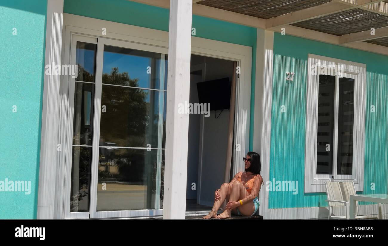 Donna rilassante, cabina turchese, libertà estiva: Una giovane donna siede tranquillamente alla porta di una rustica casa sulla spiaggia turchese, che irradia l'atmosfera delle vacanze, sl Foto Stock